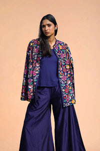Woman wearing navy palazzo pants, blue top, and floral embroidered blazer against a peach background