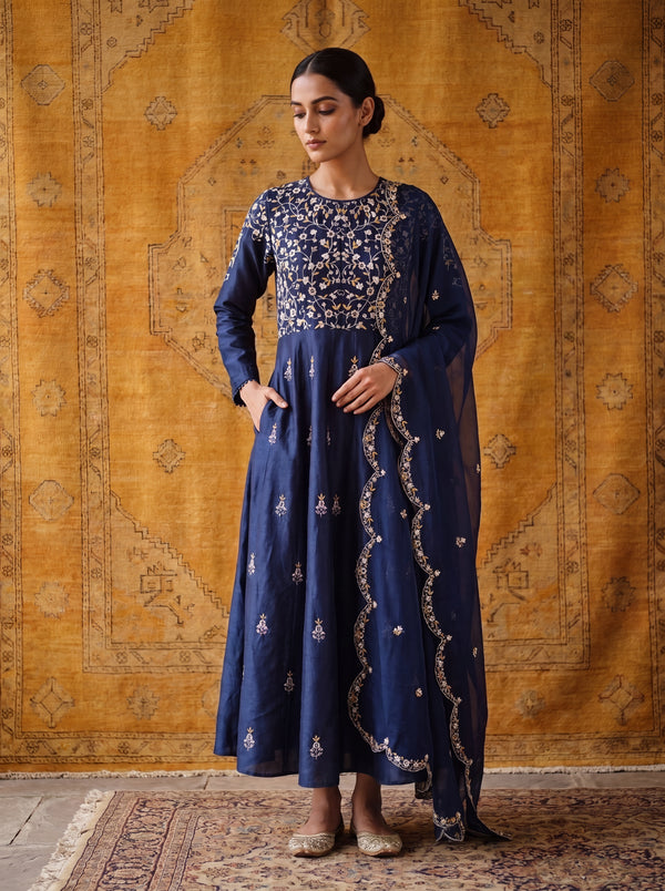 A woman models a navy blue traditional long dress with intricate gold embroidery, standing in front of a patterned golden-yellow rug.