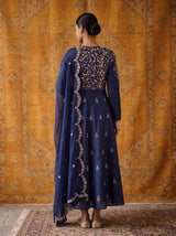 A woman stands with her back to the camera wearing a navy blue traditional outfit with intricate gold embroidery on the back and scattered gold motifs on the skirt, paired with a matching sheer dupatta. She is posed in front of a large golden patterned tapestry.