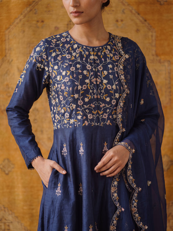 A woman wearing a navy blue embroidered traditional outfit with floral detailing in gold and beige, paired with a matching sheer dupatta, set against a warm patterned backdrop.