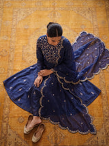 A woman sits on a large patterned rug wearing a deep navy blue traditional outfit with intricate gold embroidery, sheer dupatta, and beige flat shoes.