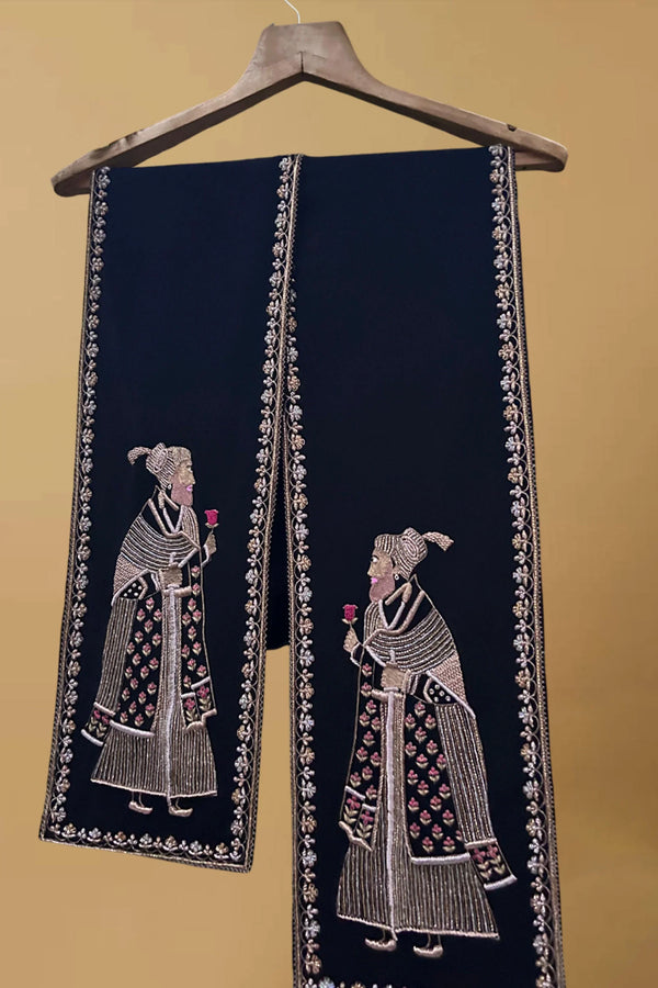Black pashmina shawl with intricate embroidered figures and floral border on hanger