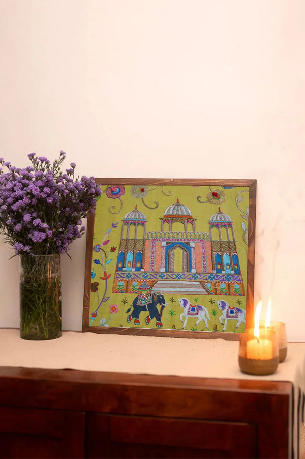 Embroidered Indian palace wall art with floral designs, elephants, and horses on a table next to purple flowers and candles.
