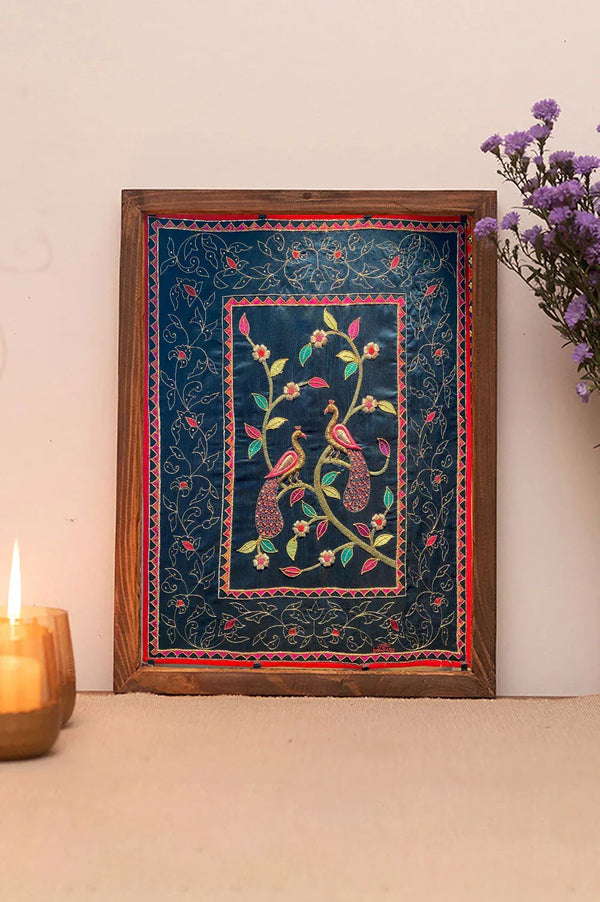 Framed embroidered peacock wall art with floral designs on blue fabric, home decor setting