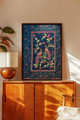Framed embroidered wall art with two peacocks and floral patterns on a blue background, displayed above a wooden cabinet in a modern living room.