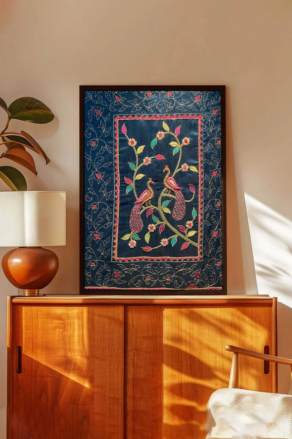 Framed embroidered wall art with two peacocks and floral patterns on a blue background, displayed above a wooden cabinet in a modern living room.